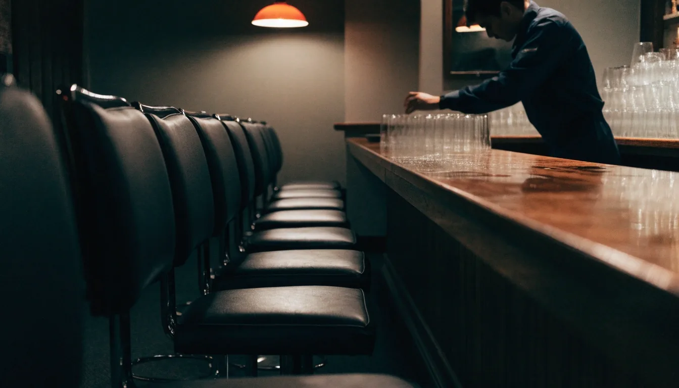 A wiped bar top and empty stools in a closed casual-dining room, lit by one amber lamp at the end of the bar
