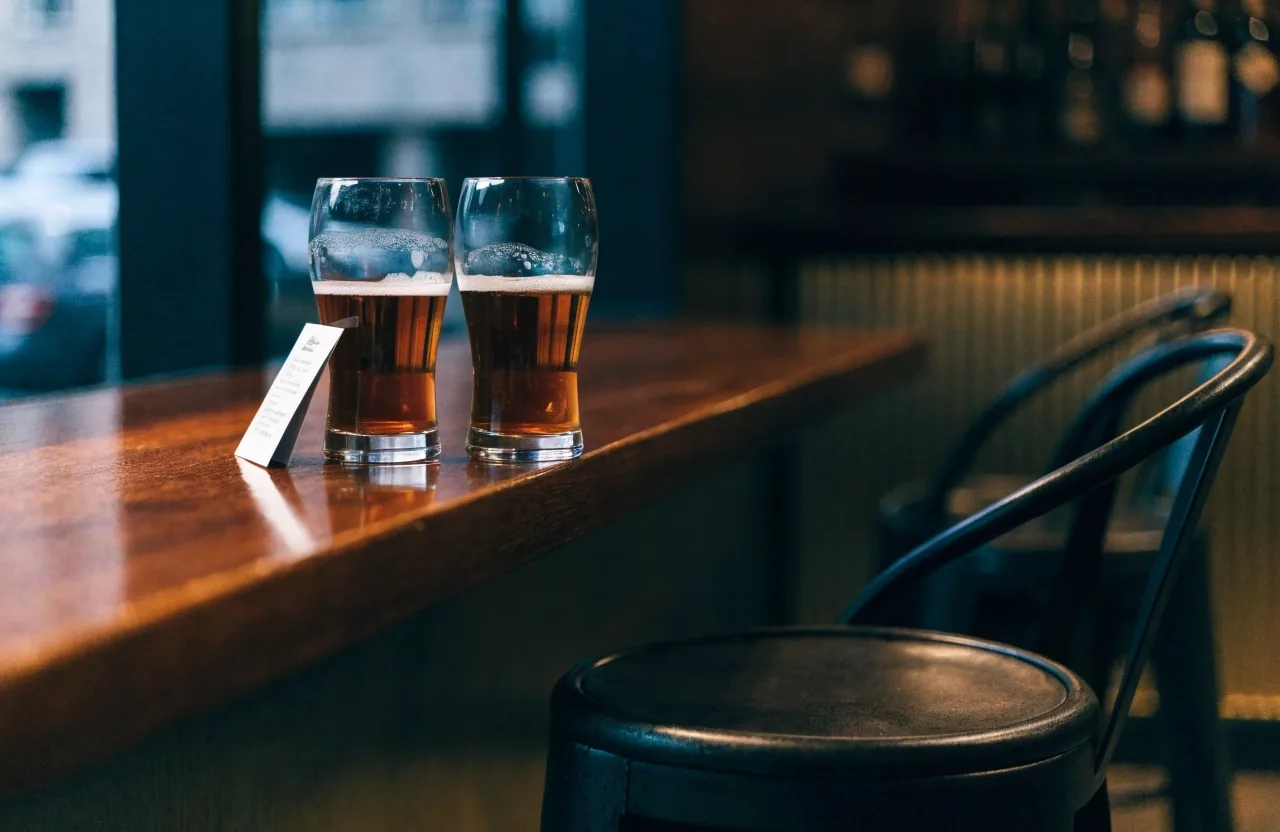 Two pours side by side on a bar table with a folded menu card between them, empty chairs behind under amber light