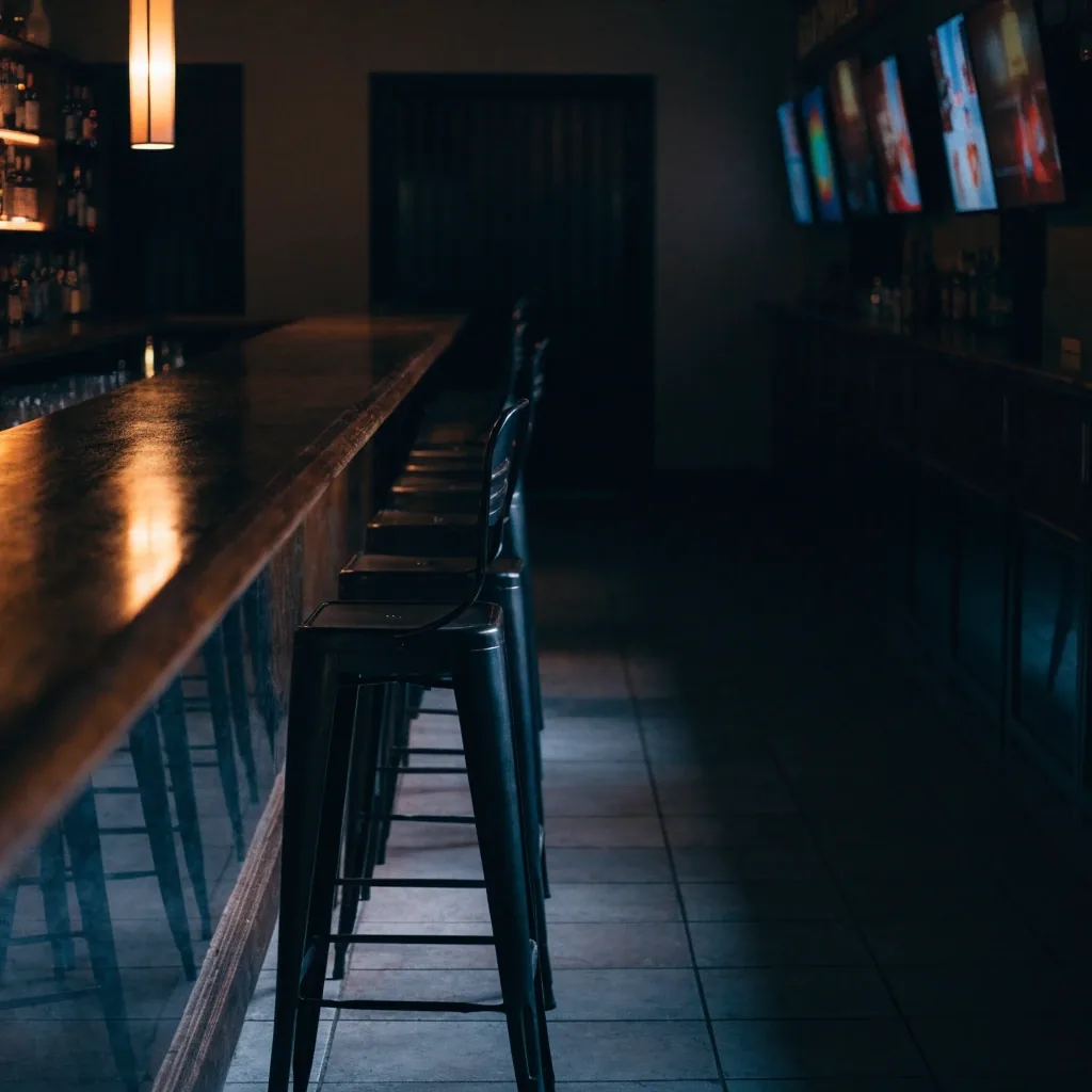 Emptied sports-bar interior at night with the counter wiped clean, stools turned up, and one amber lamp lit over the back bar