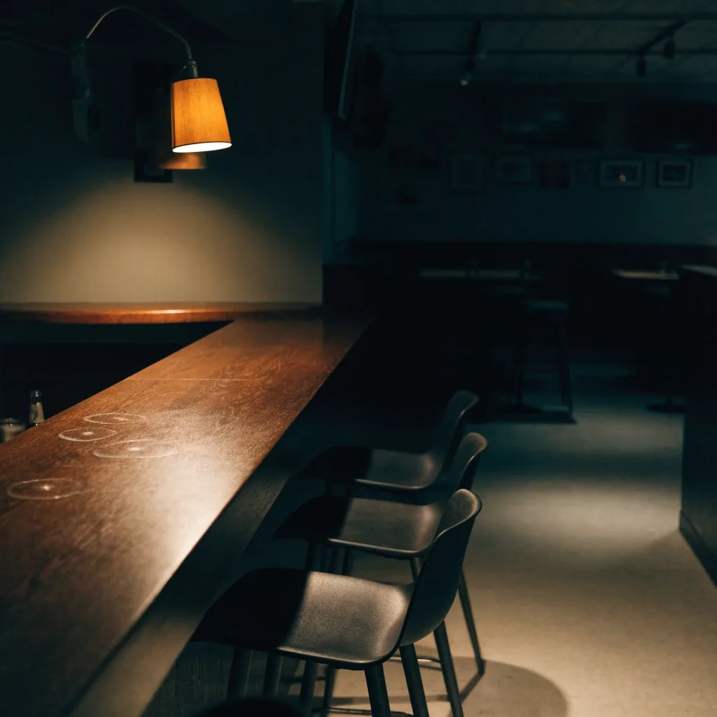 Quiet bar corner after last call with one glass ring on the wiped wood and empty stools under a single amber lamp